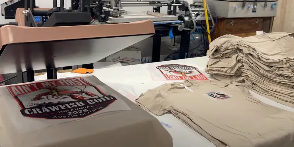 Freshly printed crawfish boil tee on the heat press beside a stack of finished tees in our Houston shop